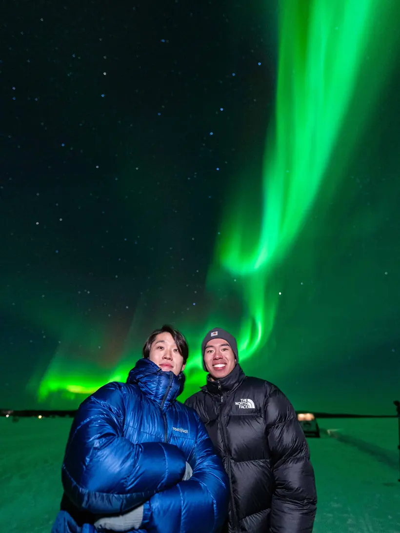 Two friends smiling under a bright green aurora curtain